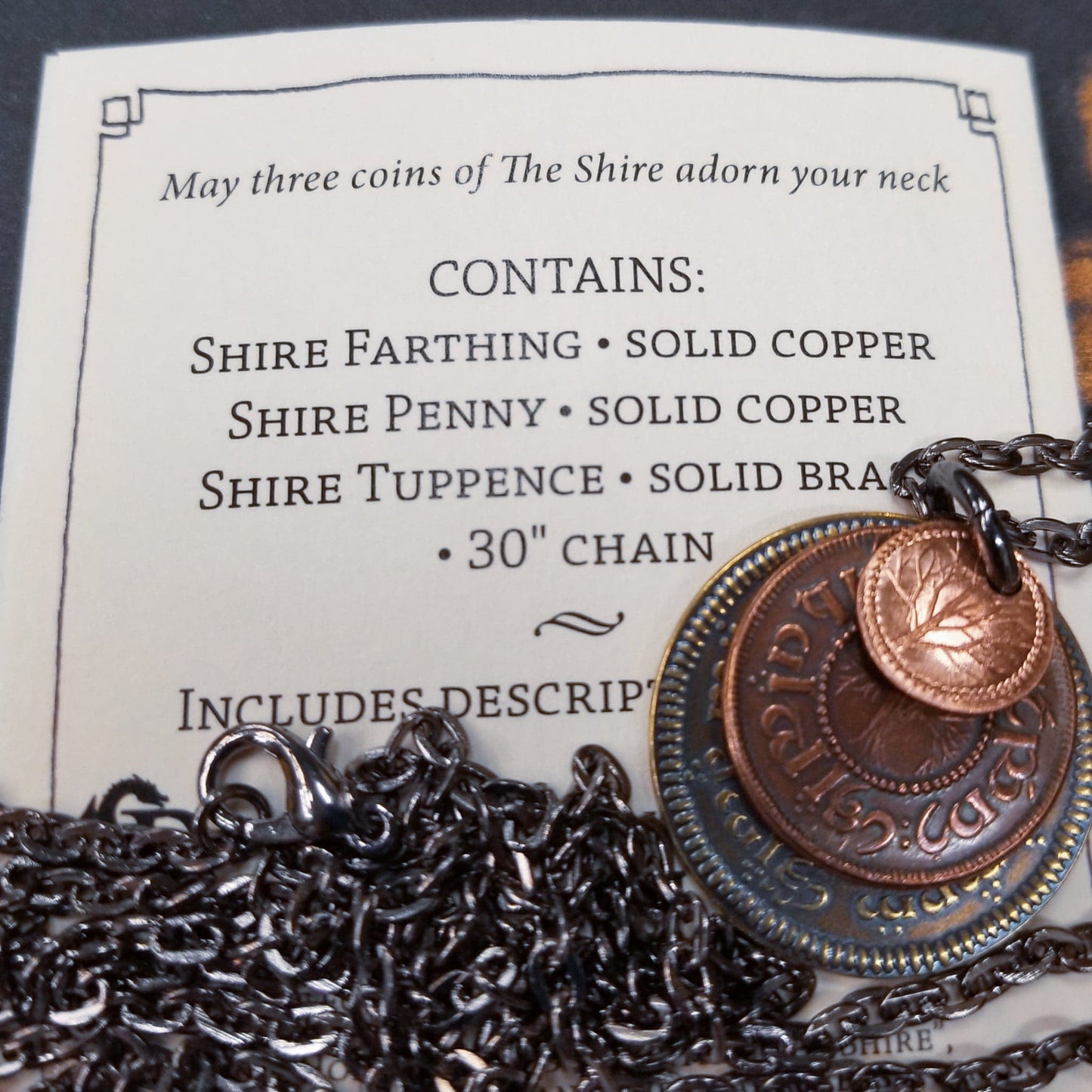 A layered necklace containing a set of three coins from the Shire in the The Lord of the Rings. A 30" chain holds three coins from The Shire coins: a brass Shire Tuppence, a copper Shire Penny, and a tiny copper Shire Farthing. Coin text is inscribed in Tengwar. The coins will each feature a tree that will be one of the following variants: pine, elm, willow, and oak. The card inside provides translation of the coin text on each coin.