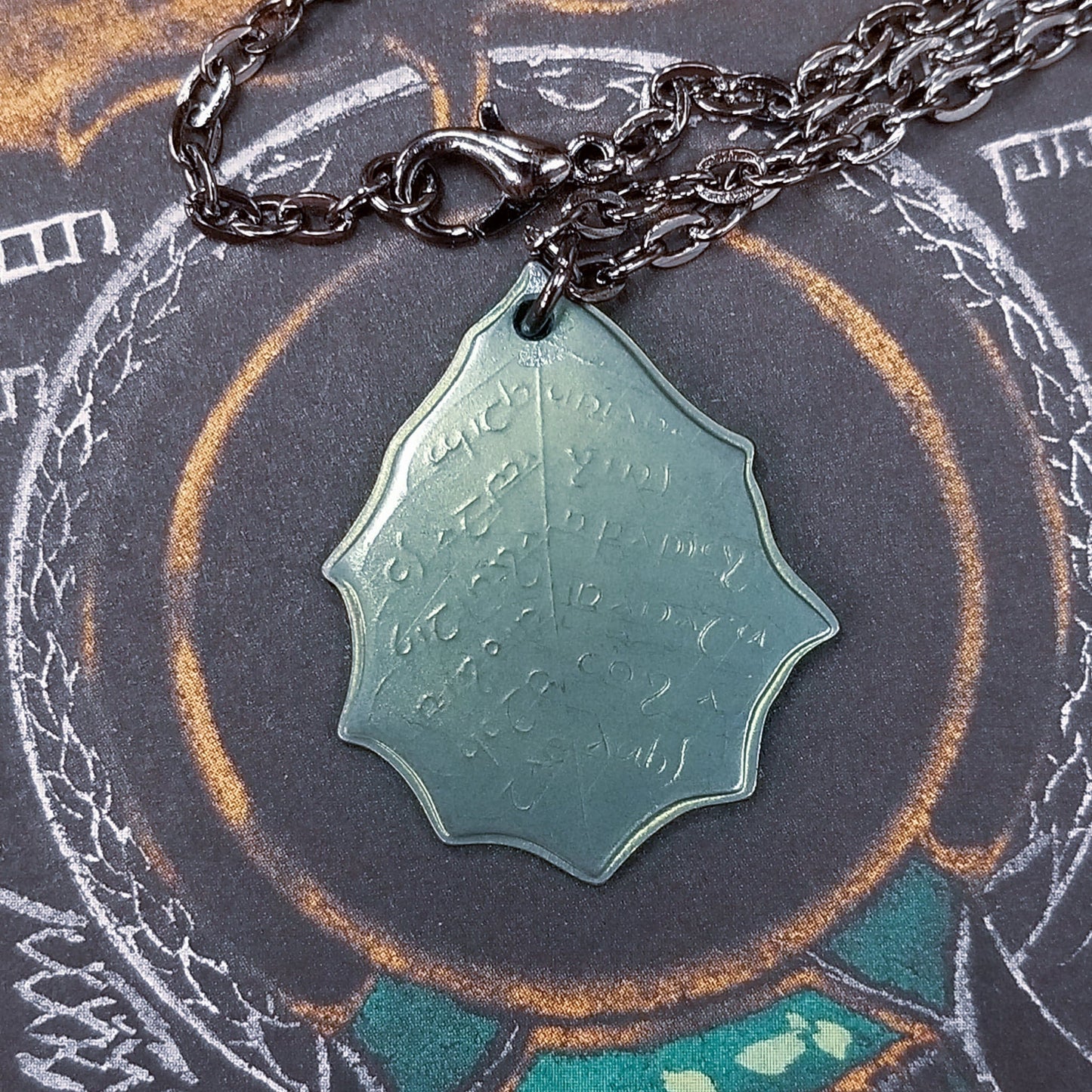 This Elvish Mithril Leaf of Spring Necklace is crafted in celebration of The Lord of the Rings by J. R. R. Tolkien. The leaf-shaped coin is struck from solid niobium, measures 2.7 cm in diameter, and weighs about 6.5 grams. Includes a 30" antique bronze cable chain with clasp. Coin artwork by Greg Franck-Weiby.  Coins are struck one at a time in the USA using antique machinery and traditional coining techniques. A colorful description is included with history, translations, and facts about the coins.