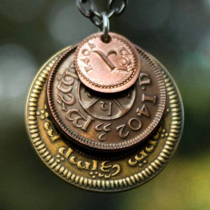 A layered necklace containing a set of three coins from the Shire in the The Lord of the Rings.  A 30" chain holds three coins from The Shire coins: a brass Shire Tuppence, a copper Shire Penny, and a tiny copper Shire Farthing. Coin text is inscribed in Tengwar. The coins will each feature a tree that will be one of the following variants: pine, elm, willow, and oak. The card inside provides translation of the coin text on each coin. 