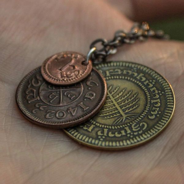 A layered necklace containing a set of three coins from the Shire in the The Lord of the Rings.  A 30" chain holds three coins from The Shire coins: a brass Shire Tuppence, a copper Shire Penny, and a tiny copper Shire Farthing. Coin text is inscribed in Tengwar. The coins will each feature a tree that will be one of the following variants: pine, elm, willow, and oak. The card inside provides translation of the coin text on each coin. 