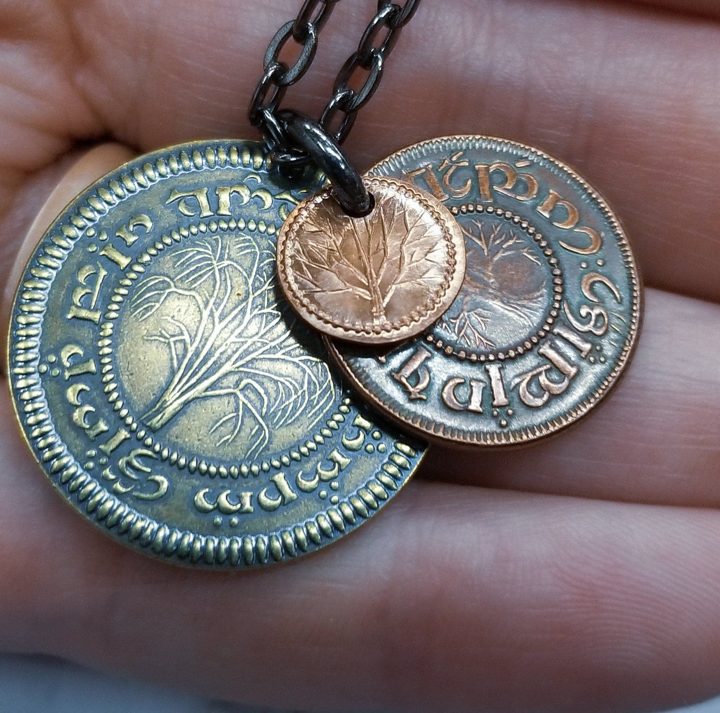 A layered necklace containing a set of three coins from the Shire in the The Lord of the Rings.  A 30" chain holds three coins from The Shire coins: a brass Shire Tuppence, a copper Shire Penny, and a tiny copper Shire Farthing. Coin text is inscribed in Tengwar. The coins will each feature a tree that will be one of the following variants: pine, elm, willow, and oak. The card inside provides translation of the coin text on each coin. 
