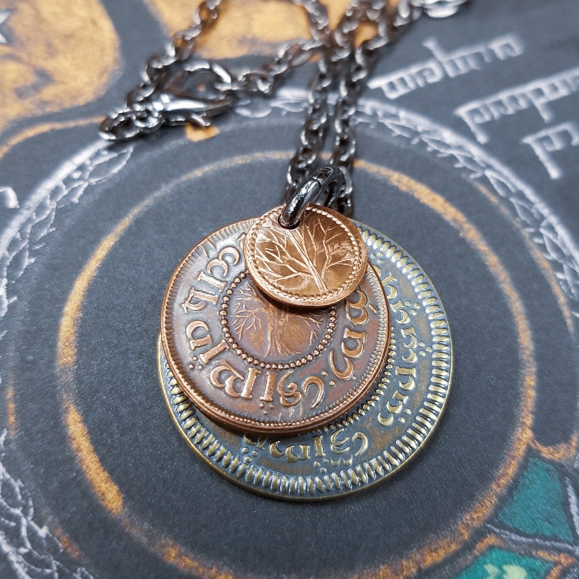 A layered necklace containing a set of three coins from the Shire in the The Lord of the Rings.  A 30" chain holds three coins from The Shire coins: a brass Shire Tuppence, a copper Shire Penny, and a tiny copper Shire Farthing. Coin text is inscribed in Tengwar. The coins will each feature a tree that will be one of the following variants: pine, elm, willow, and oak. The card inside provides translation of the coin text on each coin. 