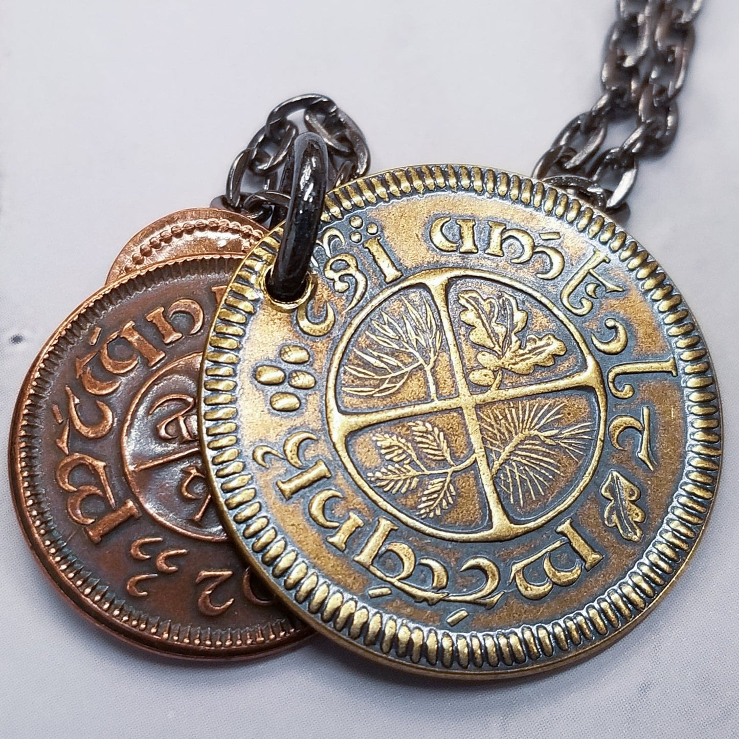 A layered necklace containing a set of three coins from the Shire in the The Lord of the Rings.  A 30" chain holds three coins from The Shire coins: a brass Shire Tuppence, a copper Shire Penny, and a tiny copper Shire Farthing. Coin text is inscribed in Tengwar. The coins will each feature a tree that will be one of the following variants: pine, elm, willow, and oak. The card inside provides translation of the coin text on each coin. 