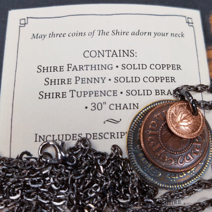 A layered necklace containing a set of three coins from the Shire in the The Lord of the Rings.  A 30" chain holds three coins from The Shire coins: a brass Shire Tuppence, a copper Shire Penny, and a tiny copper Shire Farthing. Coin text is inscribed in Tengwar. The coins will each feature a tree that will be one of the following variants: pine, elm, willow, and oak. The card inside provides translation of the coin text on each coin. 
