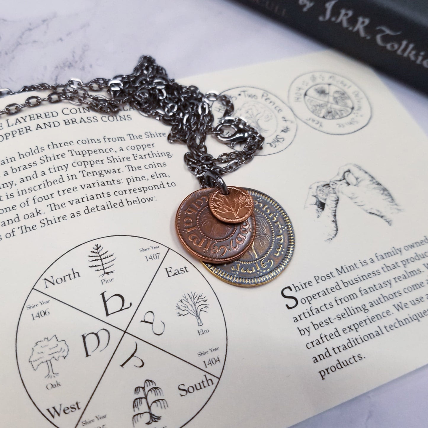 A layered necklace containing a set of three coins from the Shire in the The Lord of the Rings.  A 30" chain holds three coins from The Shire coins: a brass Shire Tuppence, a copper Shire Penny, and a tiny copper Shire Farthing. Coin text is inscribed in Tengwar. The coins will each feature a tree that will be one of the following variants: pine, elm, willow, and oak. The card inside provides translation of the coin text on each coin. 