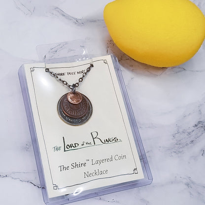 A layered necklace containing a set of three coins from the Shire in the The Lord of the Rings.  A 30" chain holds three coins from The Shire coins: a brass Shire Tuppence, a copper Shire Penny, and a tiny copper Shire Farthing. Coin text is inscribed in Tengwar. The coins will each feature a tree that will be one of the following variants: pine, elm, willow, and oak. The card inside provides translation of the coin text on each coin. 